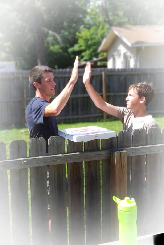 Two neighbors awkwardly high-fiving over a wooden backyard fence with an open pizza box balanced on top and a bright neon green water bottle nearby. Two neighbors awkwardly high-fiving over a wooden backyard fence with an open pizza box balanced on top and a bright neon green water bottle nearby.