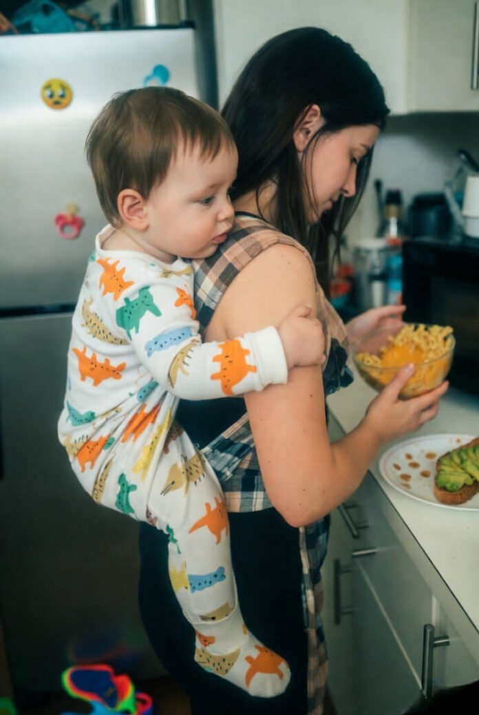 Tired mom microwaves mac and cheese with toddler in flannel carrier on her back. Tired mom microwaves mac and cheese with toddler in flannel carrier on her back.