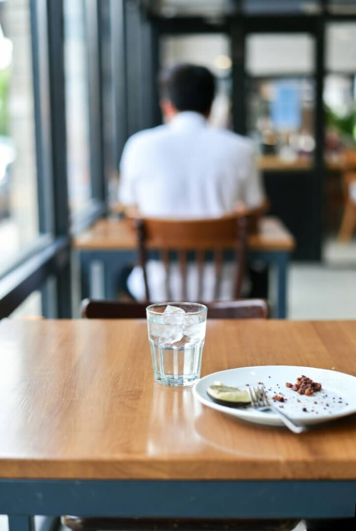 A slightly tilted candid shot of a wooden café table with an iced water glass and an empty plate of coffee grounds, someone blurred in the background. A slightly tilted candid shot of a wooden café table with an iced water glass and an empty plate of coffee grounds, someone blurred in the background.