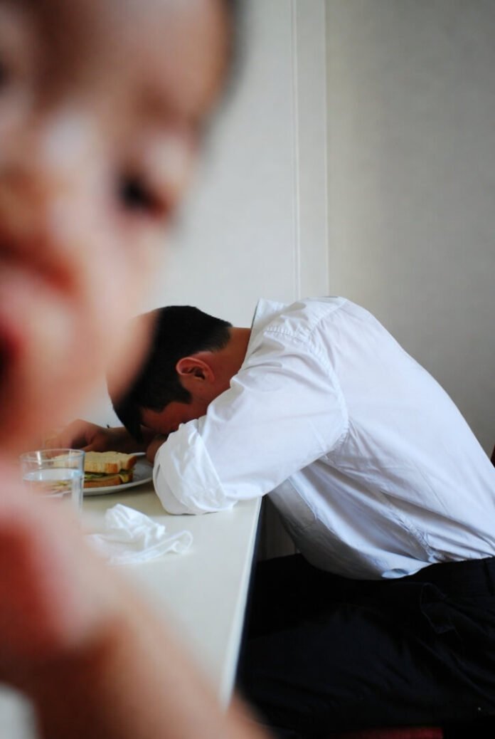 Exhausted dad slumps forward on the table, head on arms, phone nearby, shot one-handed while holding a toddler. Exhausted dad slumps forward on the table, head on arms, phone nearby, shot one-handed while holding a toddler.