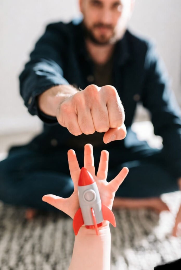 Low-angle view: child's hand bumps dad's fist holding red rocket. Low-angle view: child's hand bumps dad's fist holding red rocket.