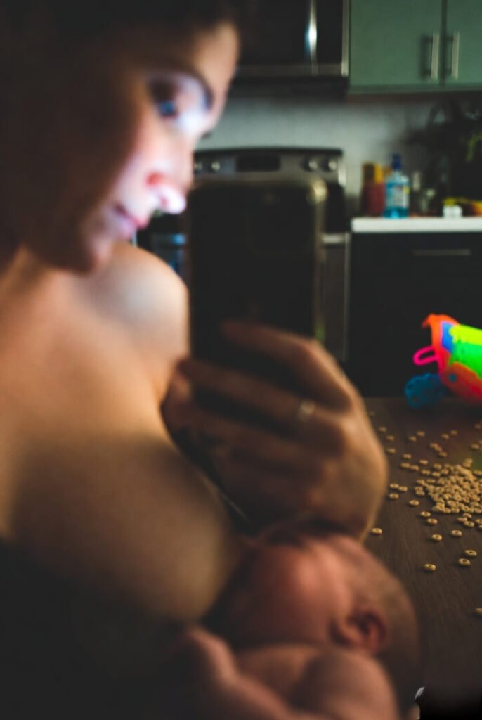 One-handed phone scroll in dim kitchen, baby latched on, Cheerios scattered below.
