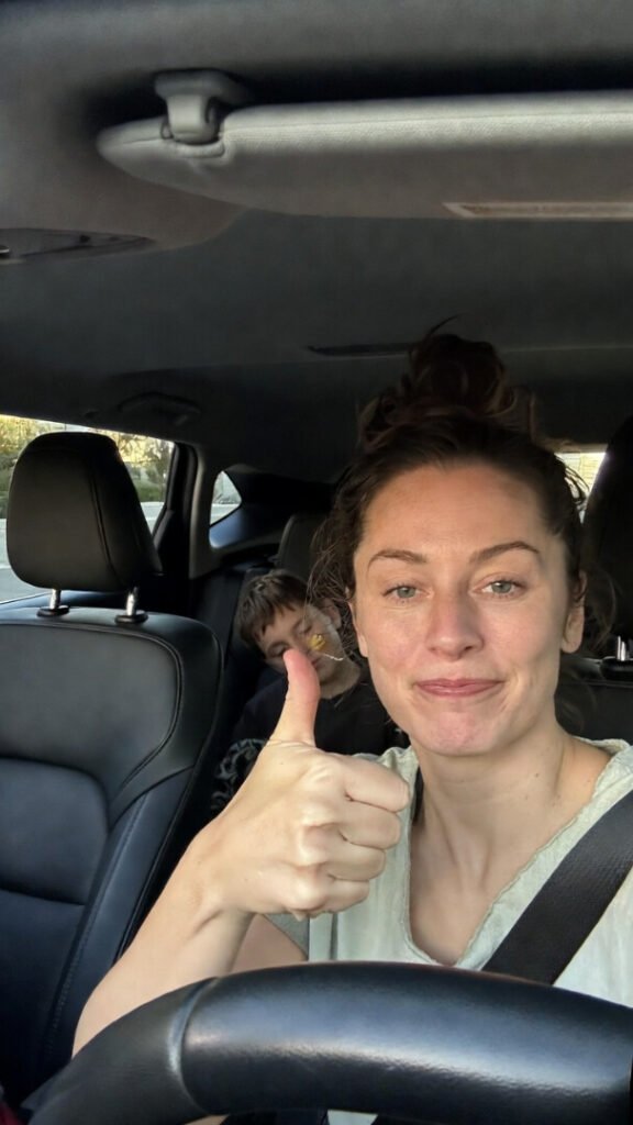 "Grainy car selfie after school pickup: tired mom with messy bun giving a forced thumbs-up in the driver's seat, young kid fast asleep in the back with fruit snack residue smeared on his cheek."