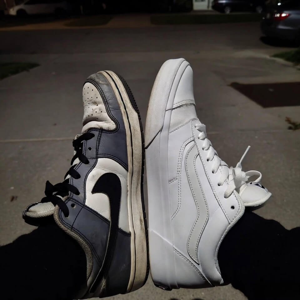 Grainy overhead shot of beat-up gray Nikes toe-to-toe with pristine white Vans on cracked concrete driveway at dusk