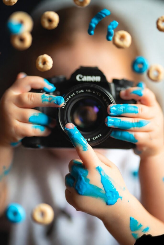 Blue paint-covered small hands, one finger suspiciously nearing lens, background of scattered Cheerios. (10 words)