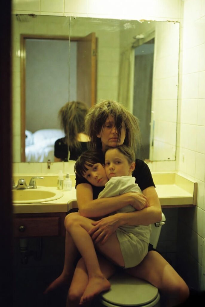 Exhausted mom on toilet lid, 8-year-old draped across lap, both dead-eyed after meltdown, wild hair.