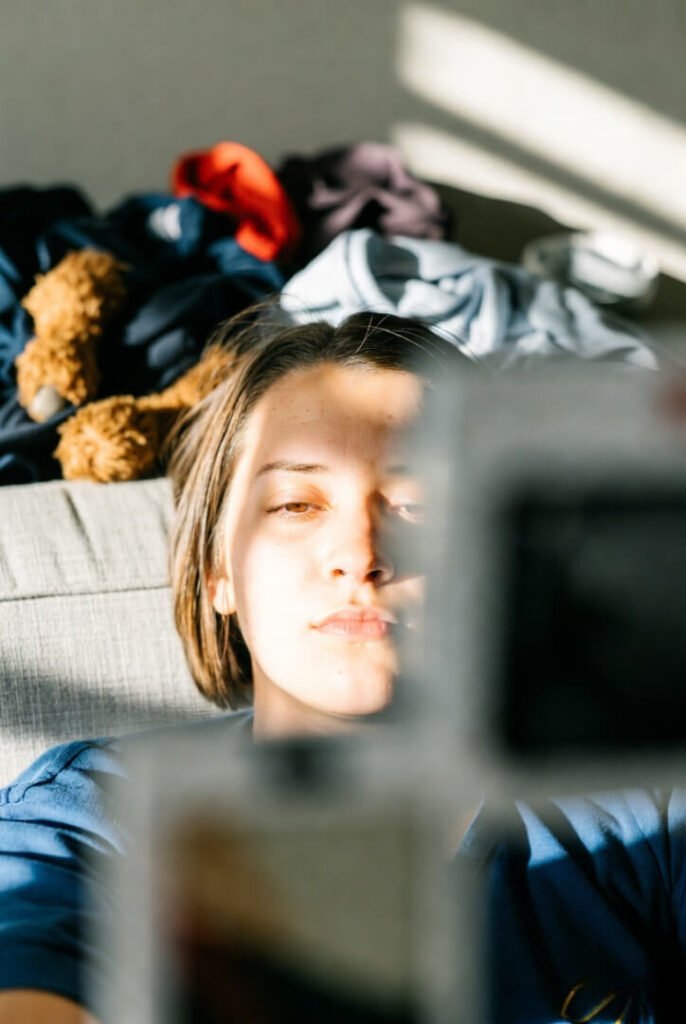 Candid half-face selfie: exhausted mom’s tired expression reflected in phone screen, messy unfolded laundry and stuffed toys piled on couch behind her.