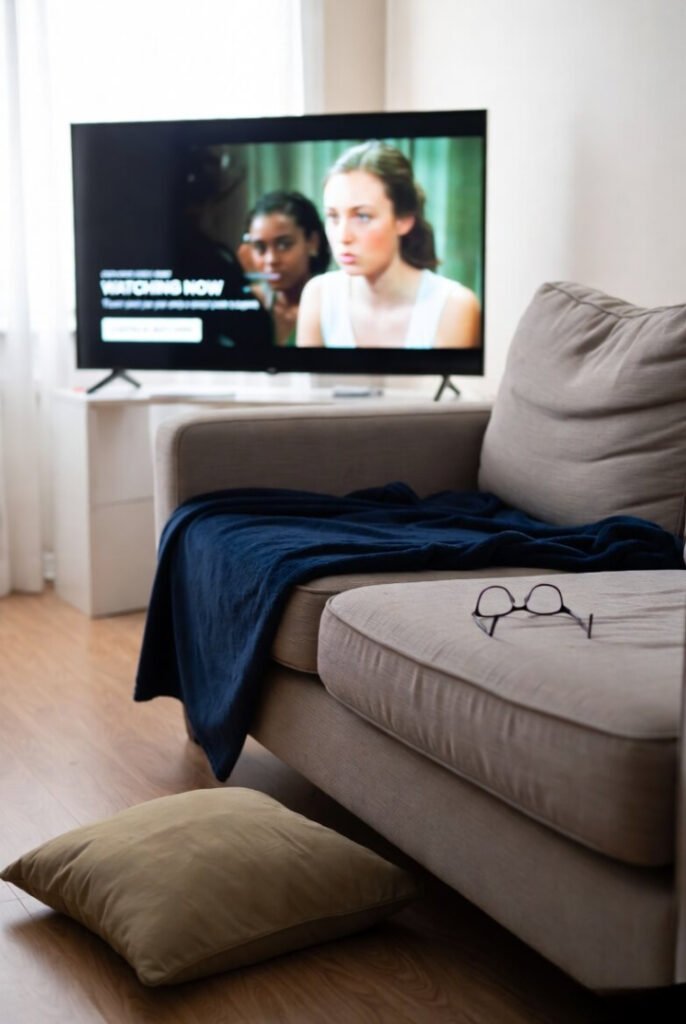 "Quiet aftermath of a fight — couch with rumpled blanket, throw pillow on the rug, reading glasses set aside, screen paused on a random teen drama still playing 'Watching Now'."