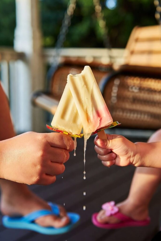 Close-up of big and tiny sibling hands sharing a melting popsicle on a porch swing, juice dripping in golden-hour light, mismatched flip-flops nearby.