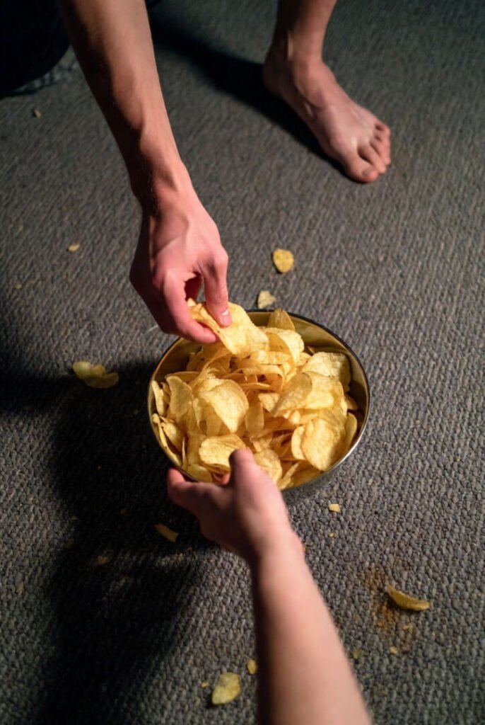 Bowl of potato chips being passed hand-to-hand at game night, messy crumbs on the floor, and a shoeless bare foot resting nearby.