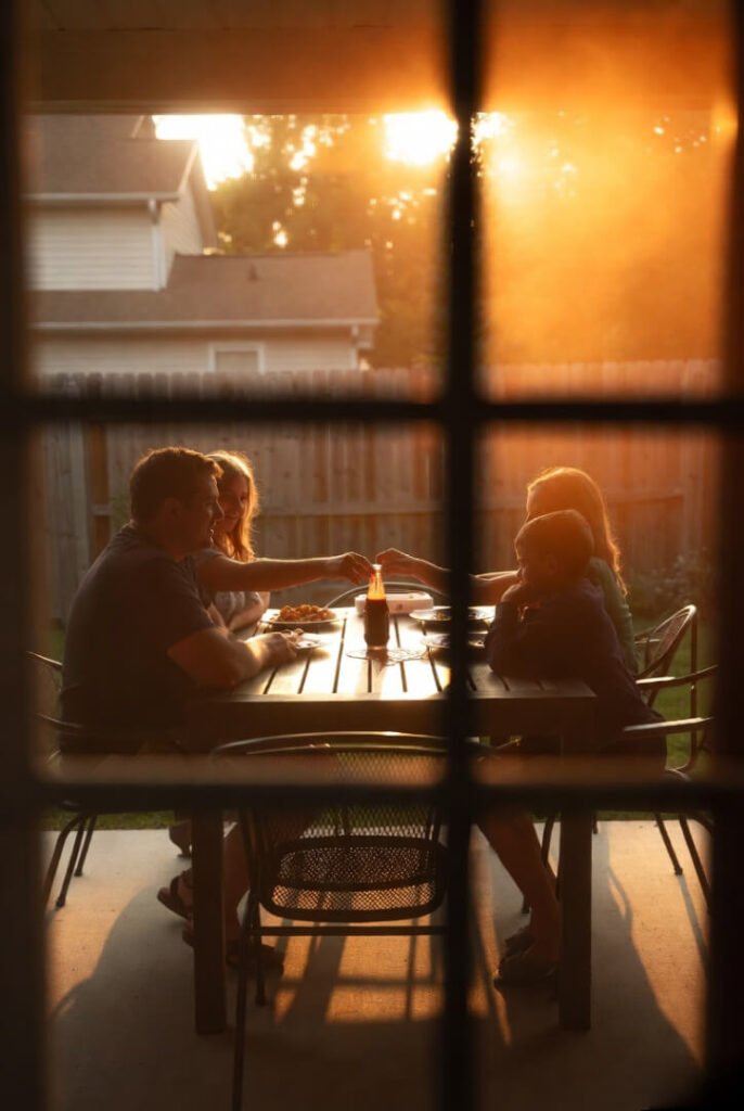 Blurry view through screen door: family at patio table in golden late-afternoon light, one empty chair, hesitant reach for ketchup bottle.