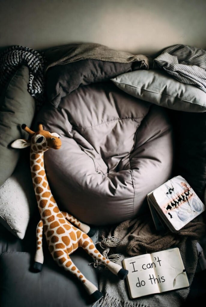 Overhead shot of failed cozy setup — messy beanbag, stuffed giraffe, gnawed board book, journal confessing “I can’t do this.”