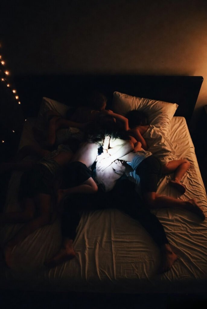 Love looks like a crime scene: exhausted parent under tangle of sleeping children, flashlight lit, foot in face.