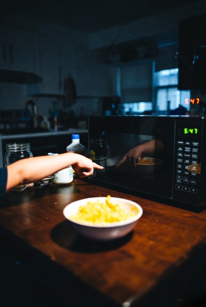 Untouched Annie’s mac & cheese cooling, child’s arm accusing across table, tired reflection in microwave door, dim evening light.